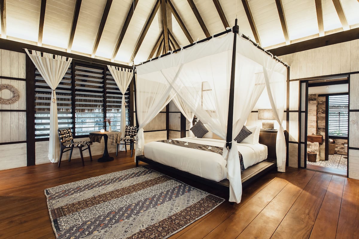 A four-poster canopy bed with a white bedspread and pillows, a patterned rug, wooden flooring, and a view of the outdoors through large windows with sheer curtains.