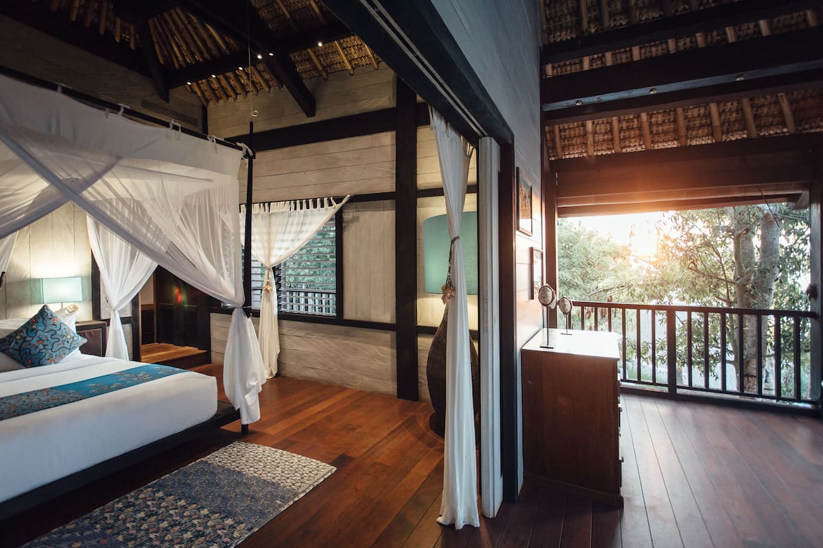 A bedroom with a large bed, a wooden floor, a balcony with a railing, and a view of trees.