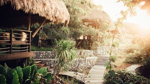 A wooden deck leading to a thatched-roof structure surrounded by lush greenery and a stone wall. - Suarga Padang Padang (Pecatu)
