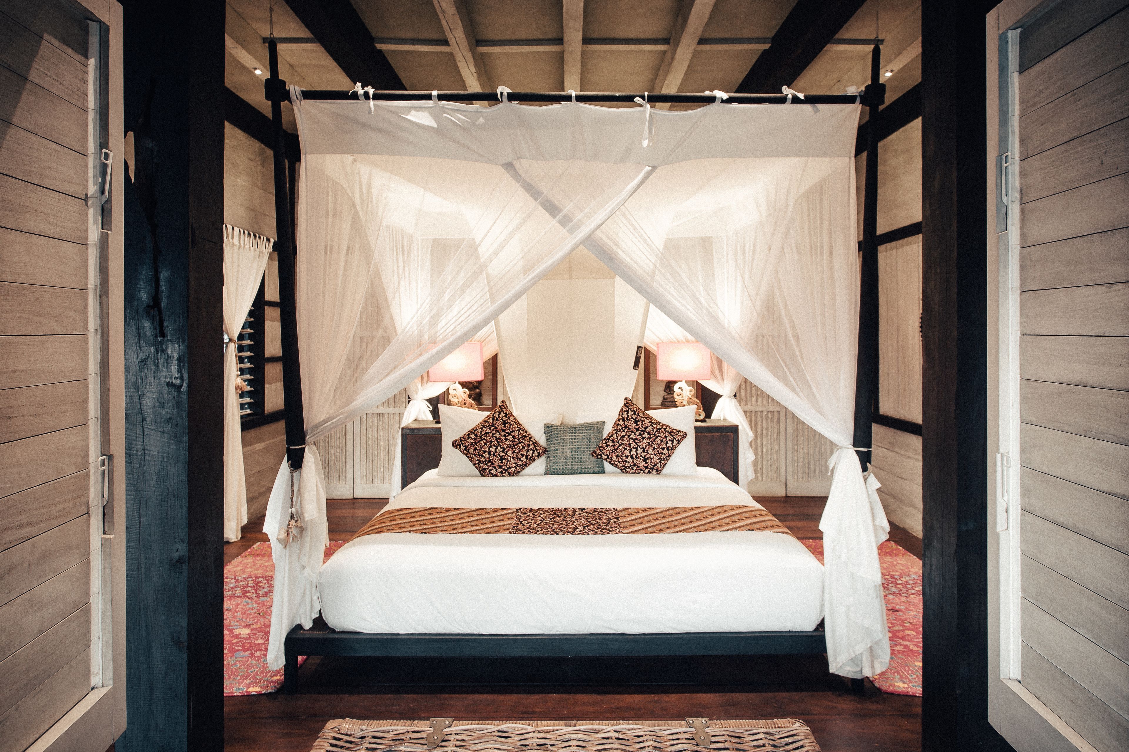 A four-poster canopy bed with white linens, two pillows, and a nightstand with a lamp.