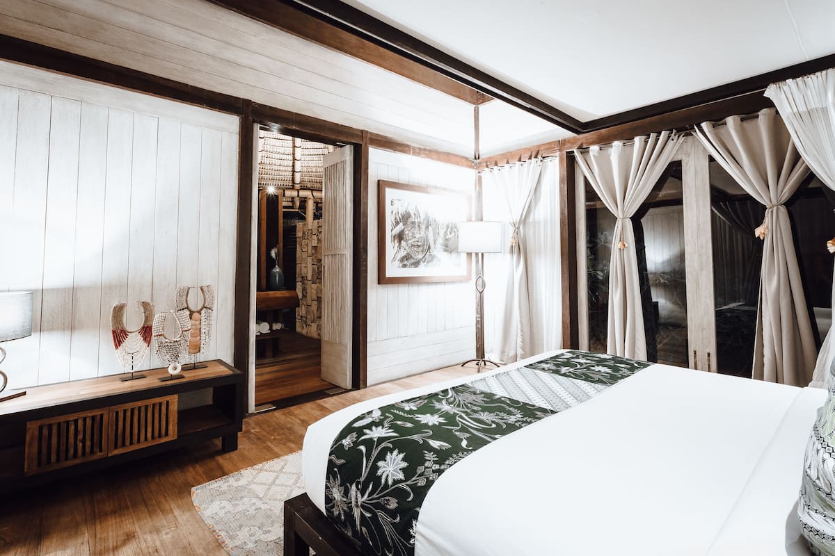 A bedroom with a wooden headboard, a bed with a green floral bedspread, a bedside table, a lamp, and a view of a hallway with a wooden door.