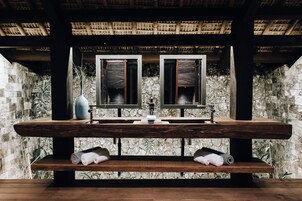 A wooden vanity with a stone wall backdrop, two mirrors, a vase, and rolled towels.