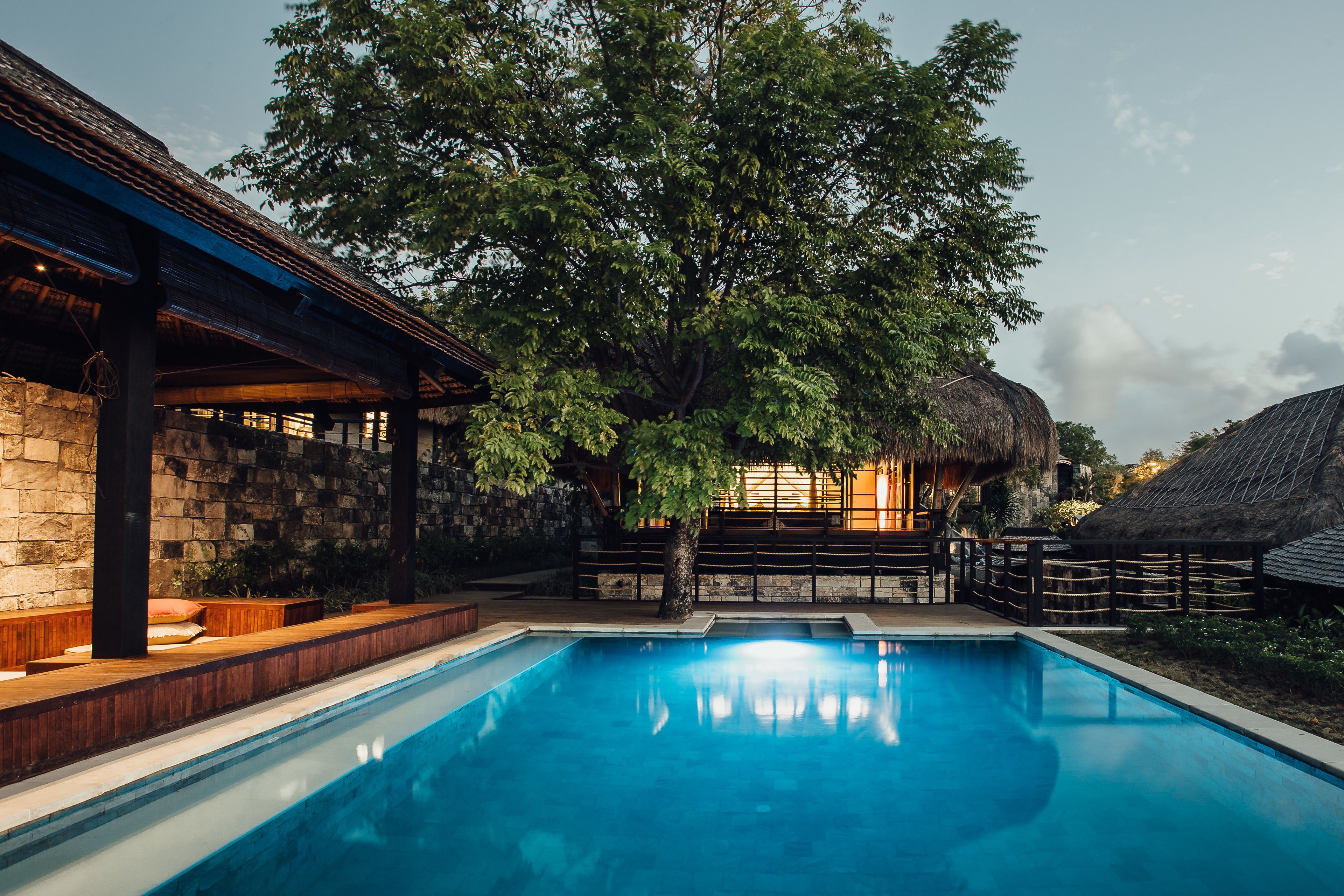 A swimming pool with a wooden deck and stone walls, surrounded by trees and a thatched-roof structure.