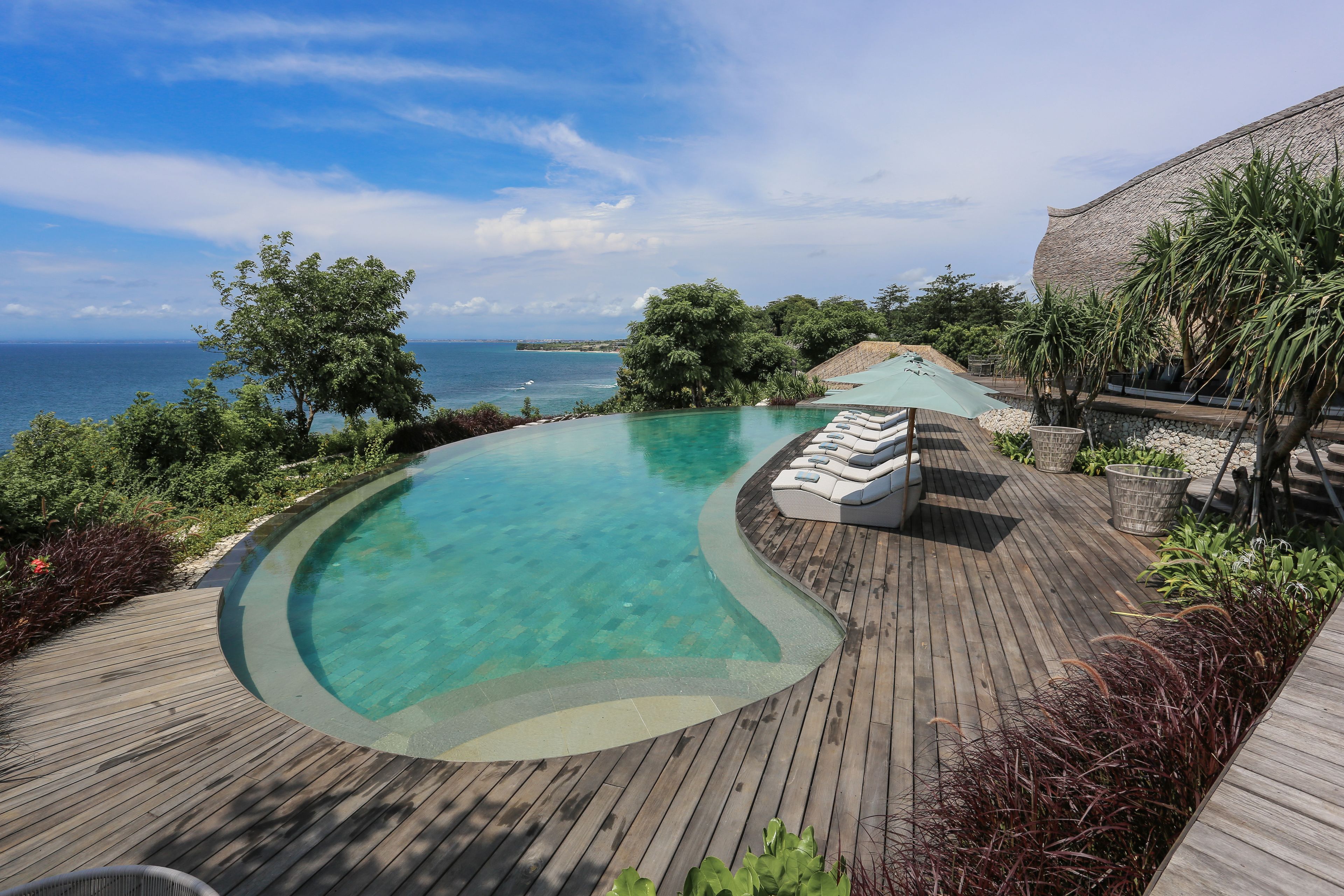 A curved infinity pool with a wooden deck, lounge chairs, and a view of the ocean.