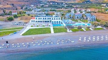 An aerial view of a coastal hotel with multiple buildings, a swimming pool, and a beach area with sun loungers.
