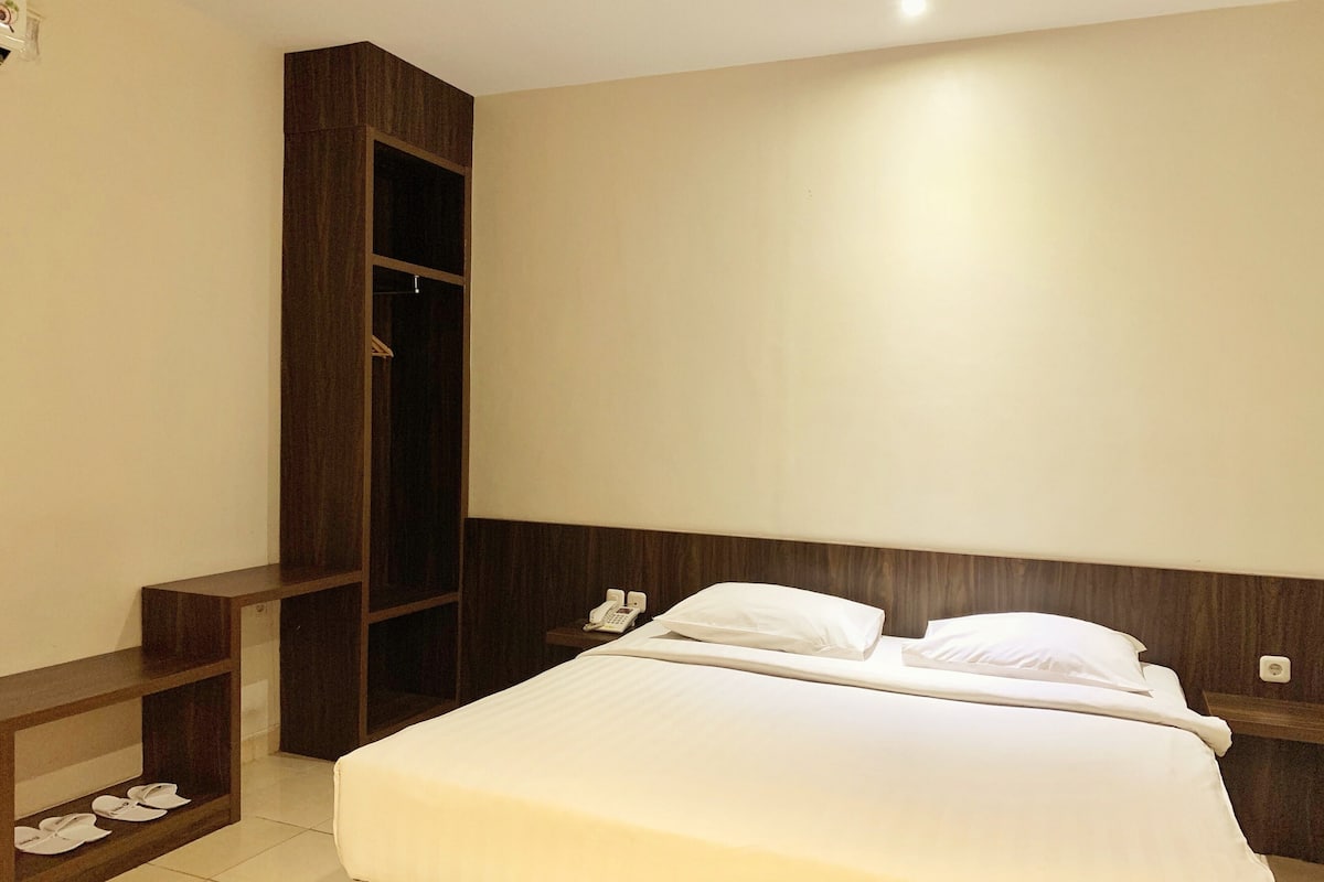A neatly arranged hotel room with a bed, a bedside telephone, a small shelf with shoes, and a wardrobe.