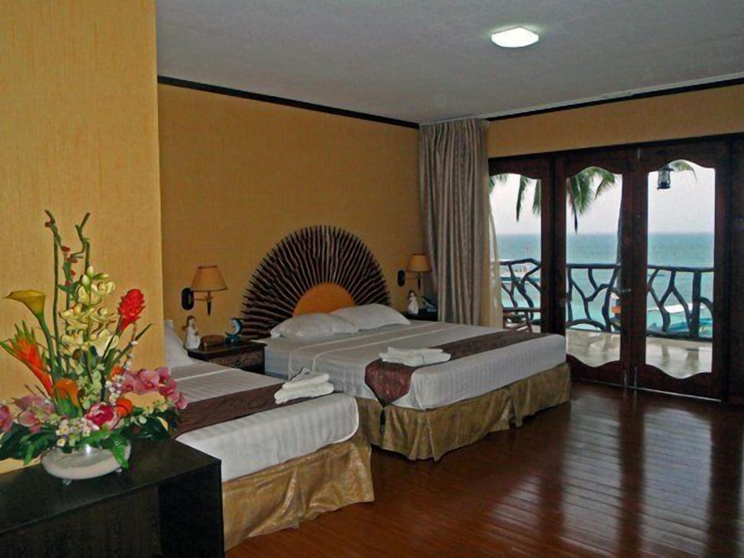 A hotel room with two beds, a large window with a view of the ocean, and a balcony with a railing.