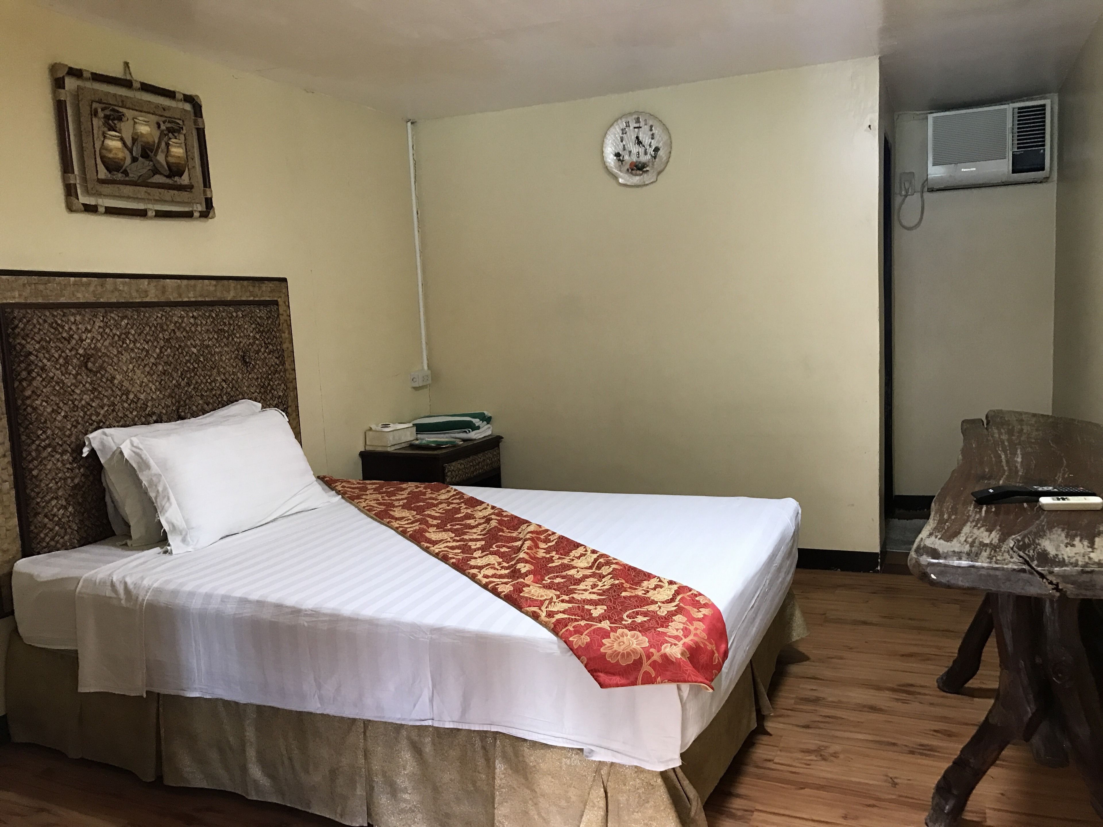 A hotel room with a bed, a nightstand, a wall clock, and an air conditioning unit.
