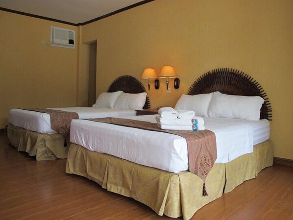 A hotel room with two beds, beige walls, wooden flooring, and a wall-mounted air conditioning unit.