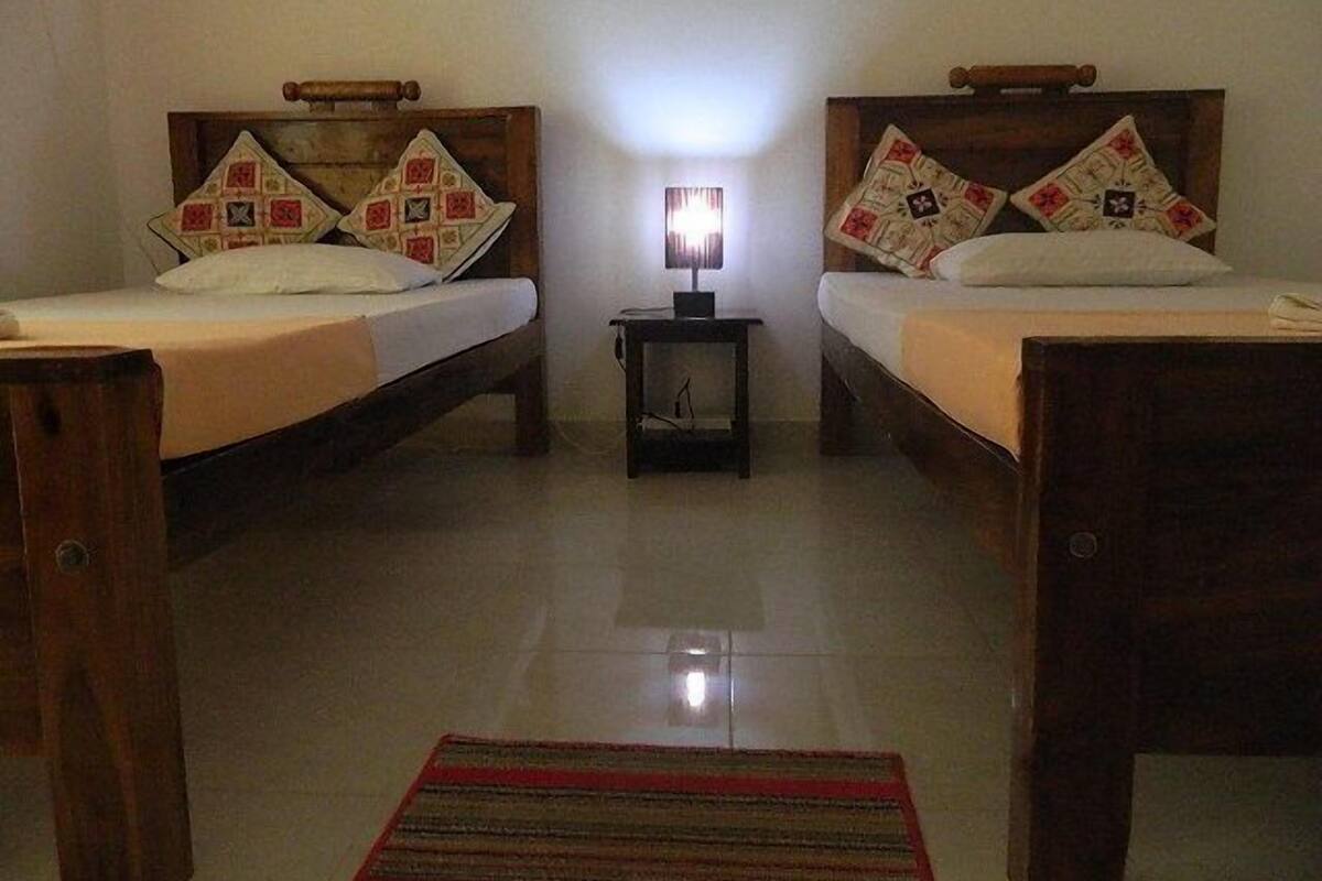 Une chambre avec deux lits, chacun doté d’une tête de lit en bois et d’un oreiller à motifs. Une petite table avec une lampe se trouve entre les lits, et un tapis rayé recouvre le sol.
