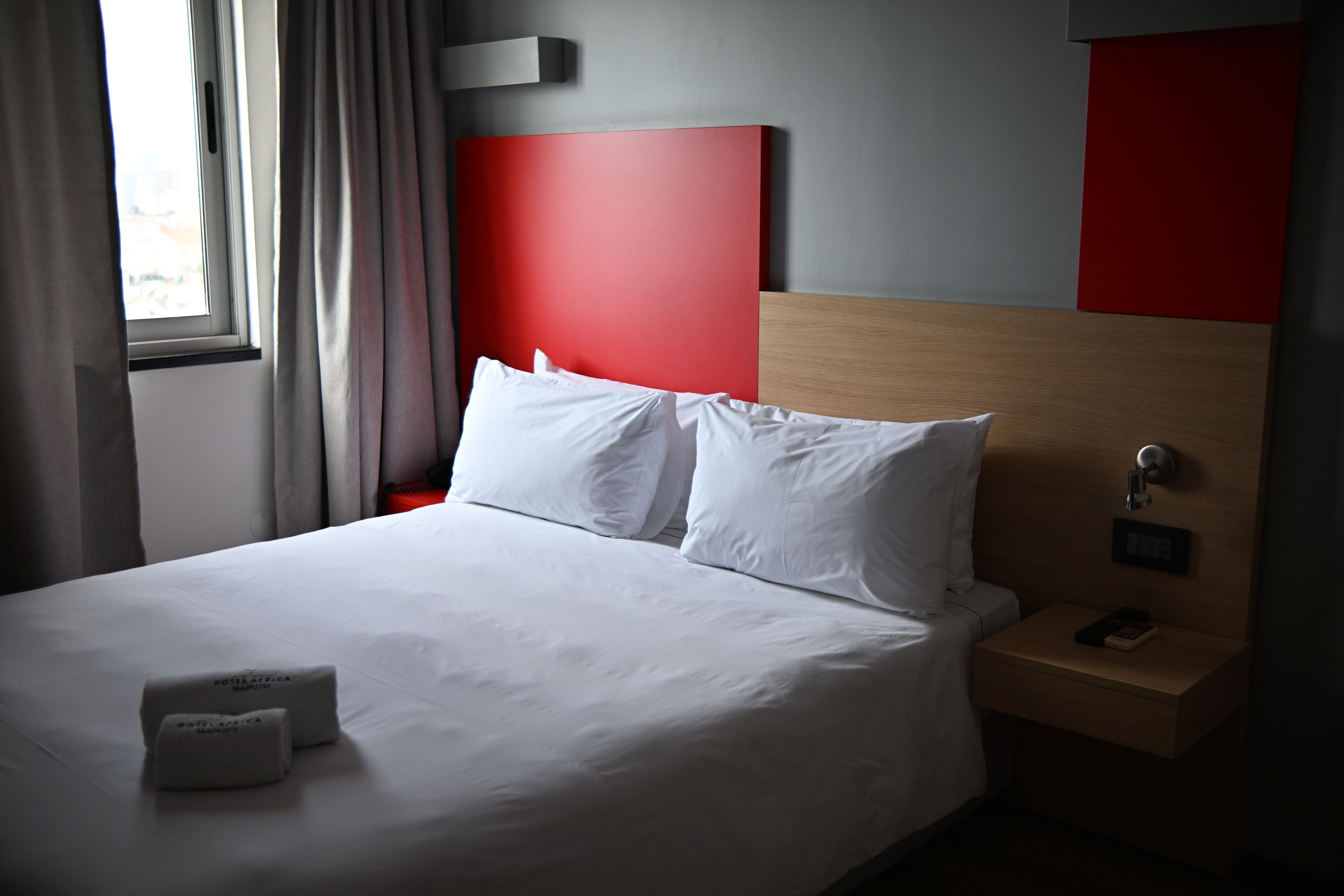 Una habitación de hotel con cama, almohadas, mesita de noche, teléfono y ventana con cortinas.