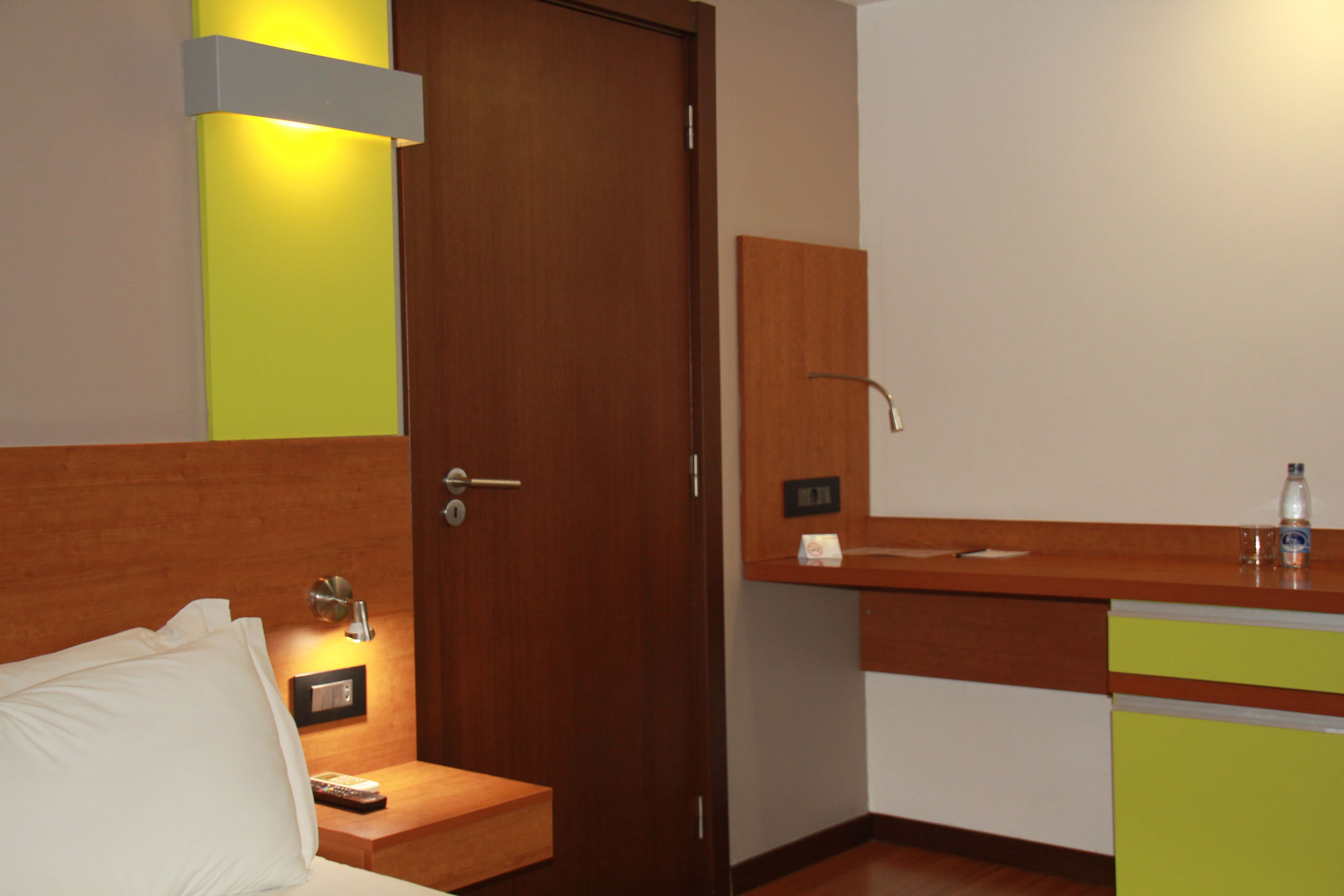 Ein Hotelzimmer mit Holztür, Nachttisch mit Lampe, kleiner Ablagefläche und einem grünen Schrank.