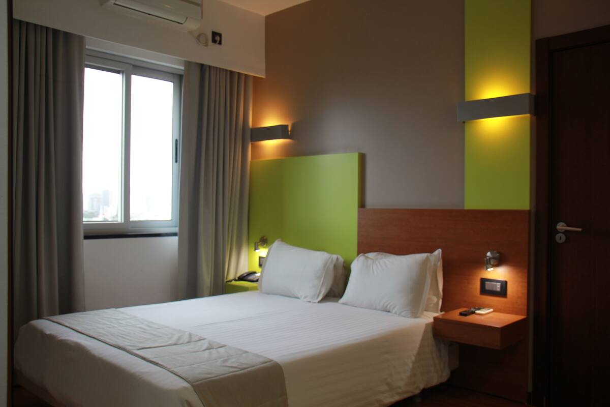 A hotel room with a large bed, a nightstand, a lamp, and a window with curtains.