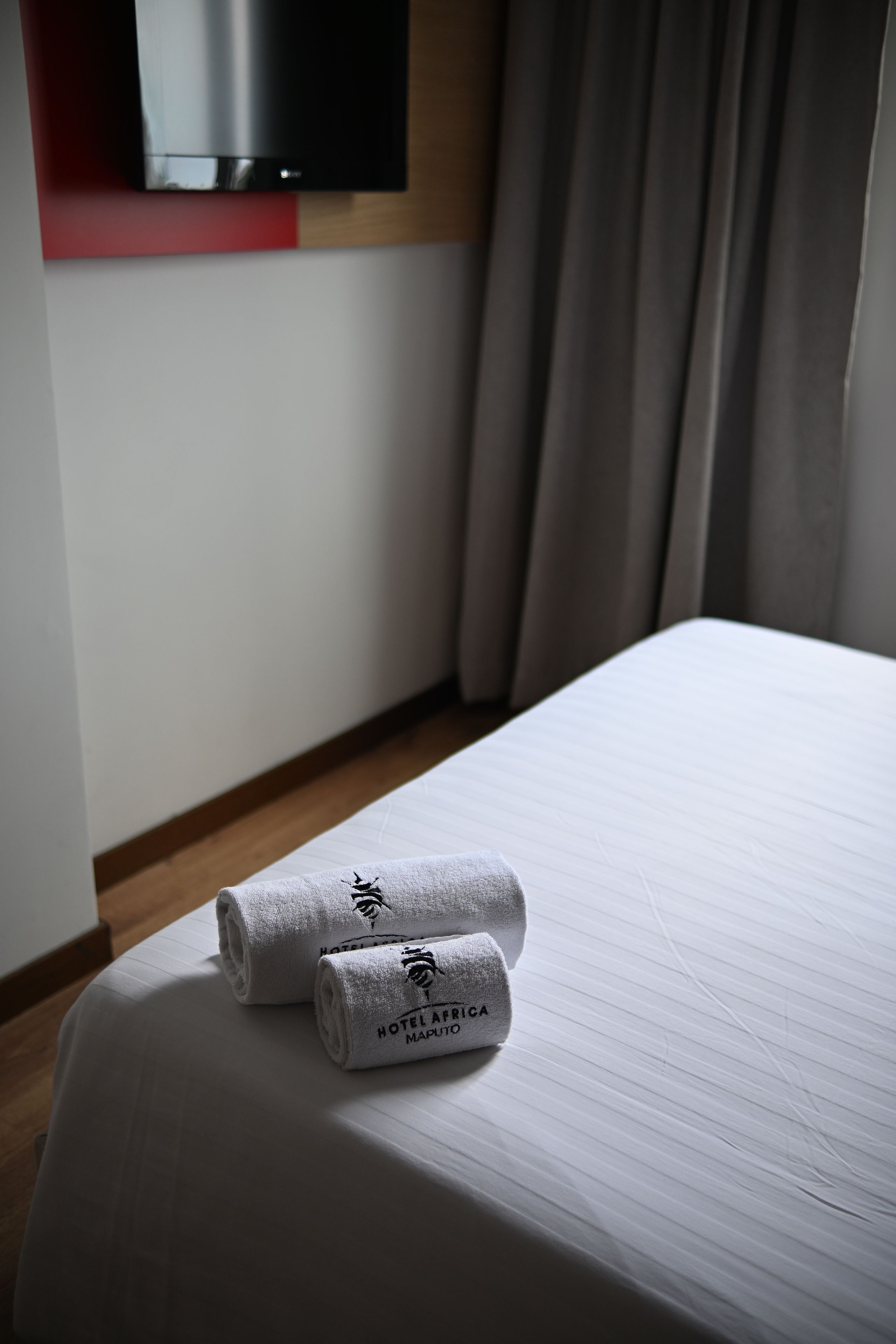 Ein Hotelzimmer mit einem ordentlich bezogenen Bett, zwei mit einem Logo versehenen gerollten Handtüchern, einem an der Wand montierten Flachbildfernseher und einem Holzboden.