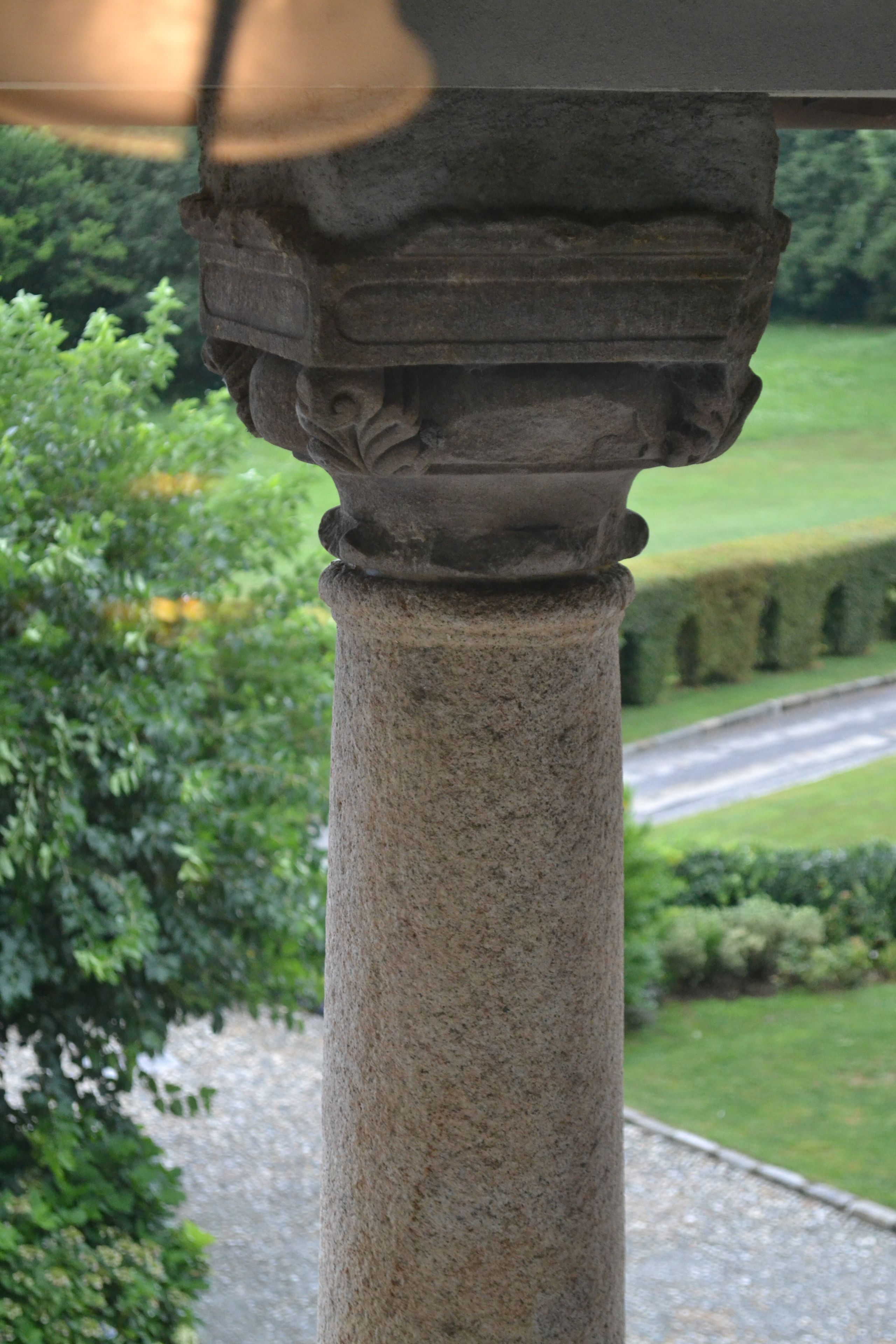 A stone column with intricate carvings, set against a backdrop of a well-manicured garden and a road.