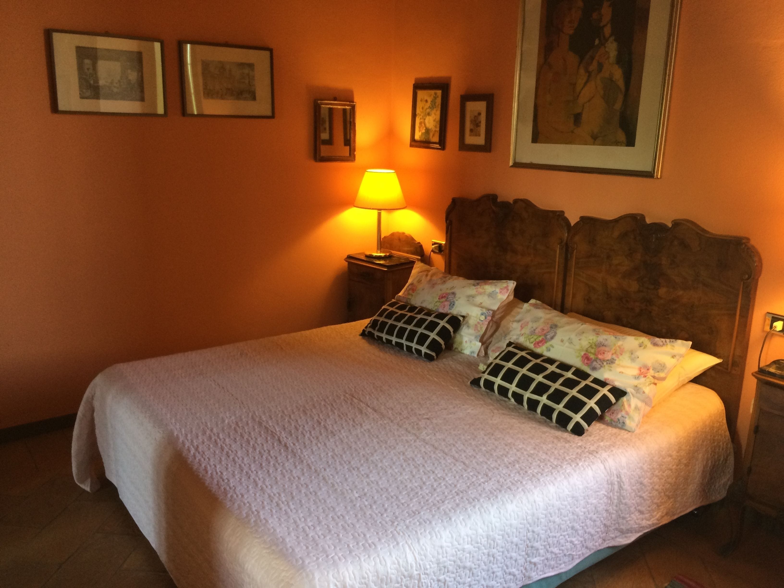 Ein ordentlich bezogenes Bett mit Blumendekoration und zwei Zierkissen, eine Nachttischlampe sowie gerahmte Bilder an der Wand.