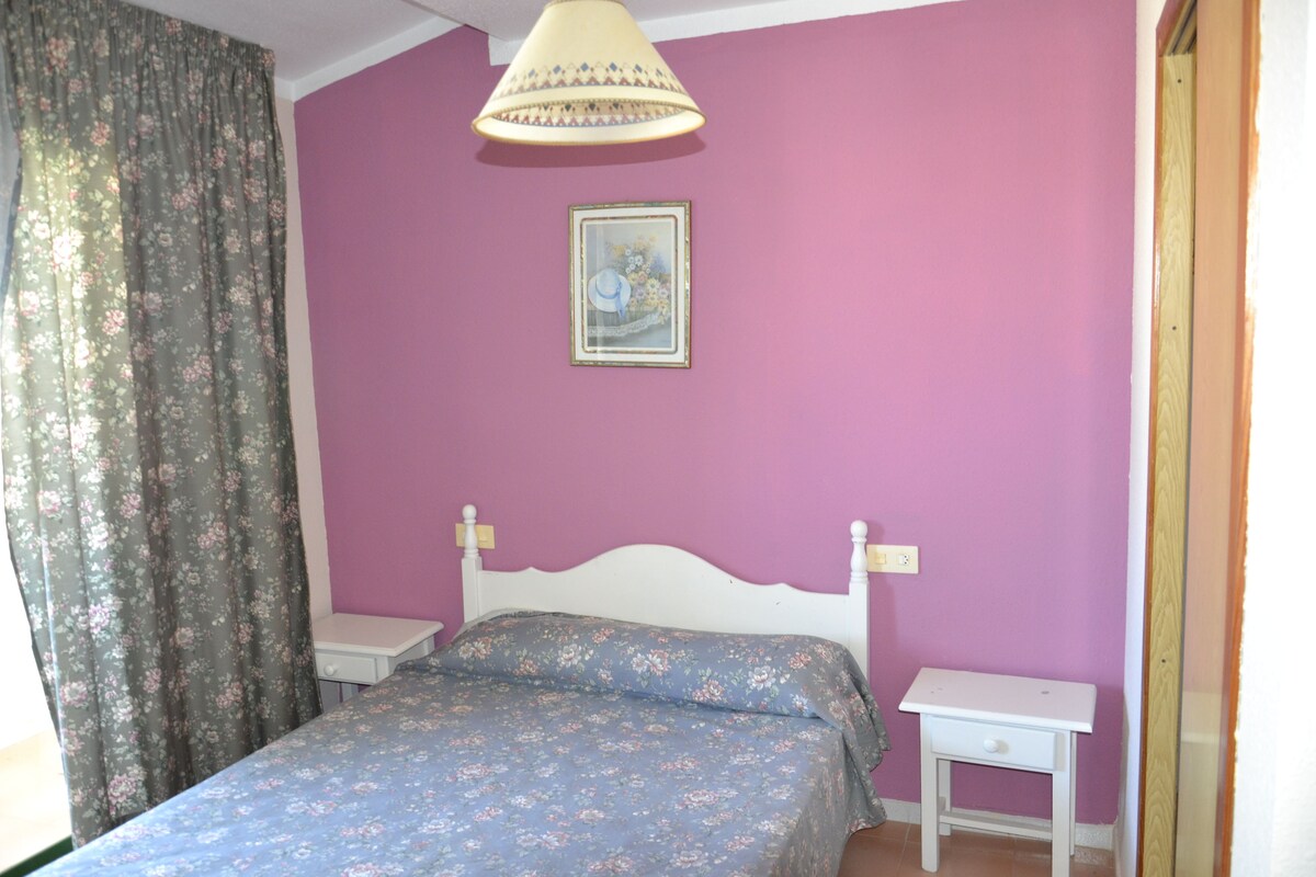 Un dormitorio con cama, mesita de noche, lámpara de pared y un cuadro enmarcado.