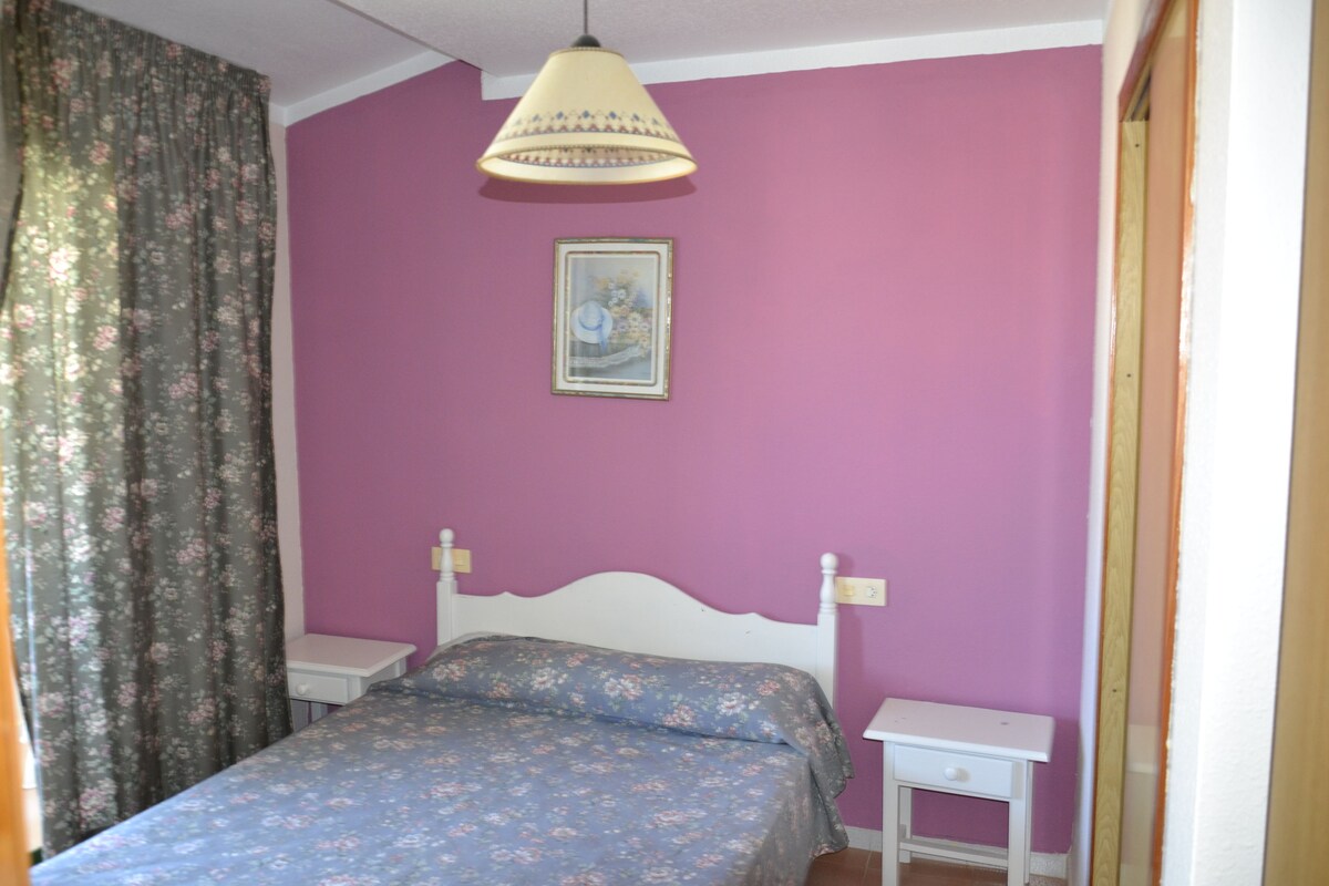 Un dormitorio con cama, mesita de noche, lámpara de pared y un cuadro enmarcado.