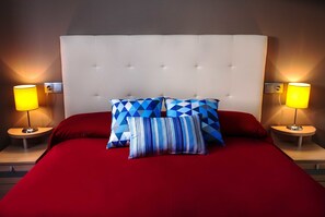 Una cama con cabecera capitonada, dos lámparas de noche y varios almohadones estampados.