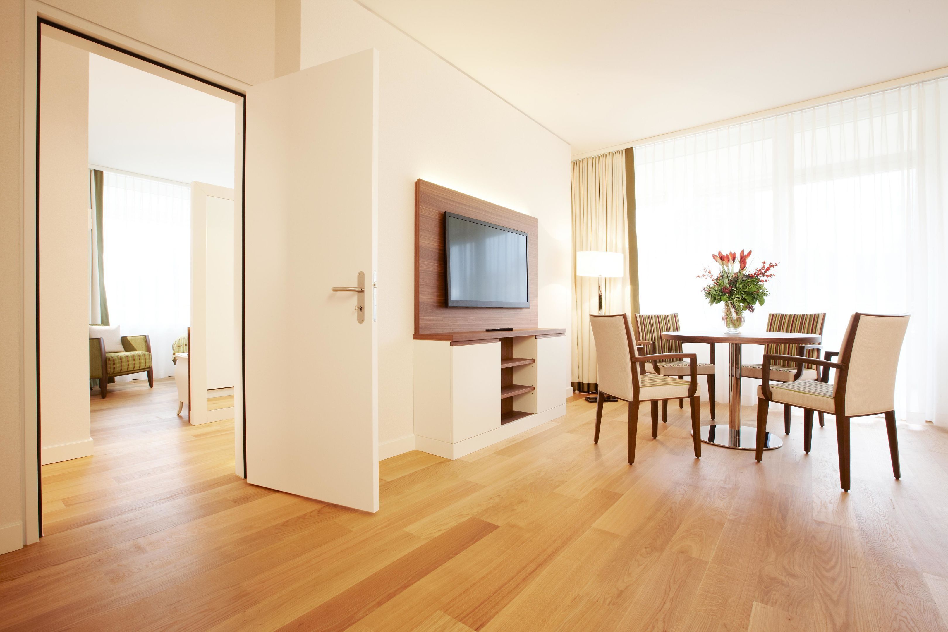 Una camera d'albergo con pavimento in legno, una televisione a schermo piatto montata su un mobile in legno, un'area pranzo con tavolo e sedie, e una finestra con tende.