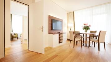 Una camera d'albergo con pavimento in legno, una televisione a schermo piatto montata su un mobile in legno, un'area pranzo con tavolo e sedie, e una finestra con tende.