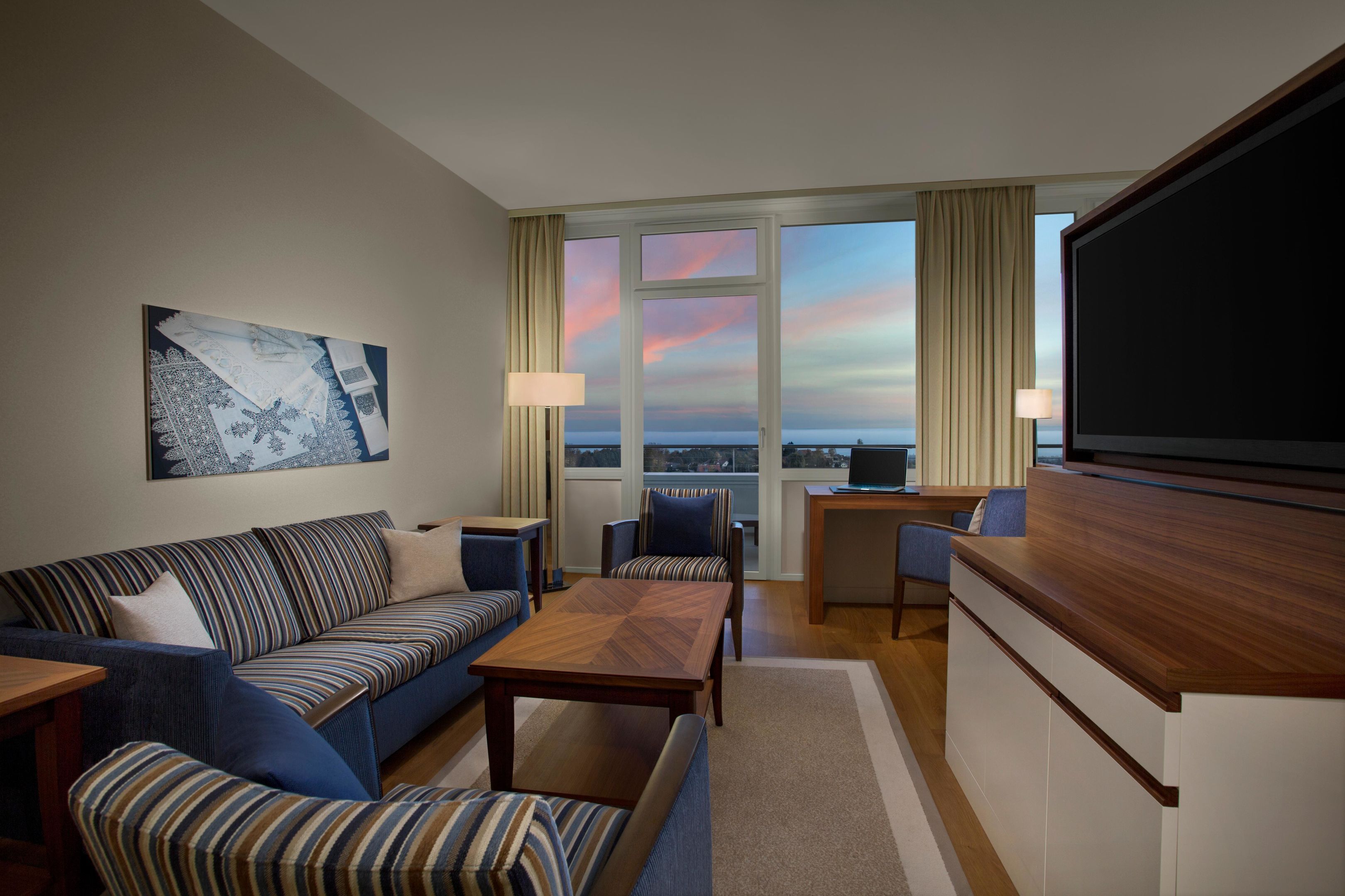 Ein modernes Hotelzimmer mit einer Couch, Sesseln, einem Couchtisch, einem Fernseher und einem großen Fenster mit Blick auf die Stadt.