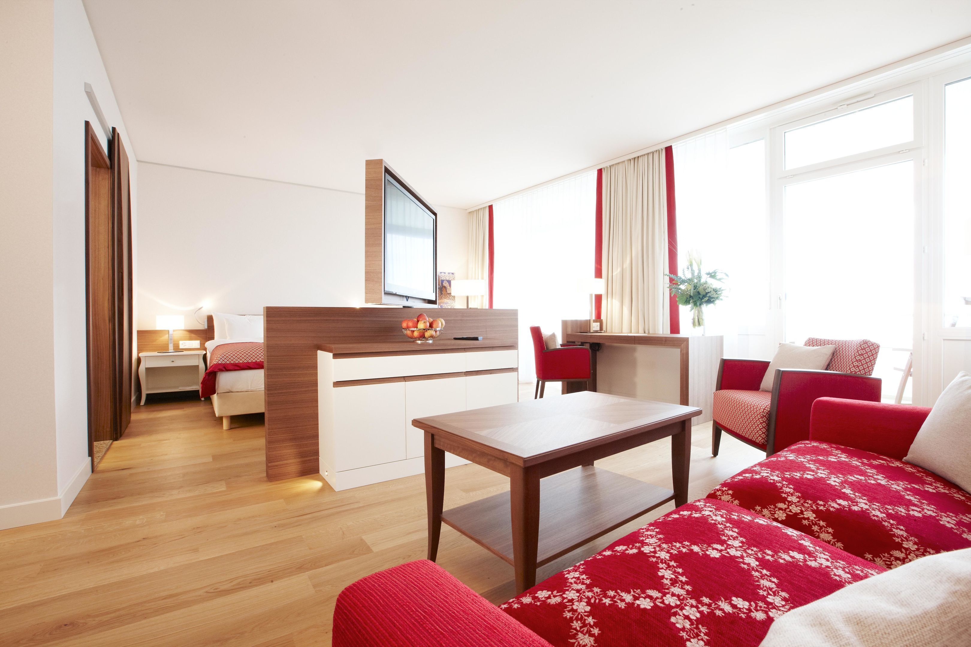 A modern hotel room with a wooden floor, a dining area, a bed with red bedding, a TV, and a window with curtains.