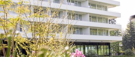 A modern multi-story building with balconies and large windows, surrounded by a well-maintained garden with various plants and flowers.