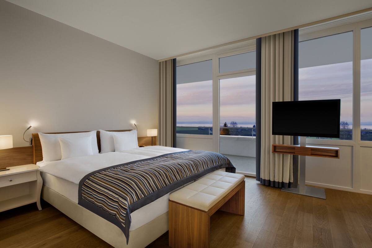 Une chambre d’hôtel moderne dotée d’un grand lit, d’un téléviseur à écran plat fixé au mur, d’une table de chevet avec une lampe et offrant une vue sur la ville grâce à de grandes fenêtres.