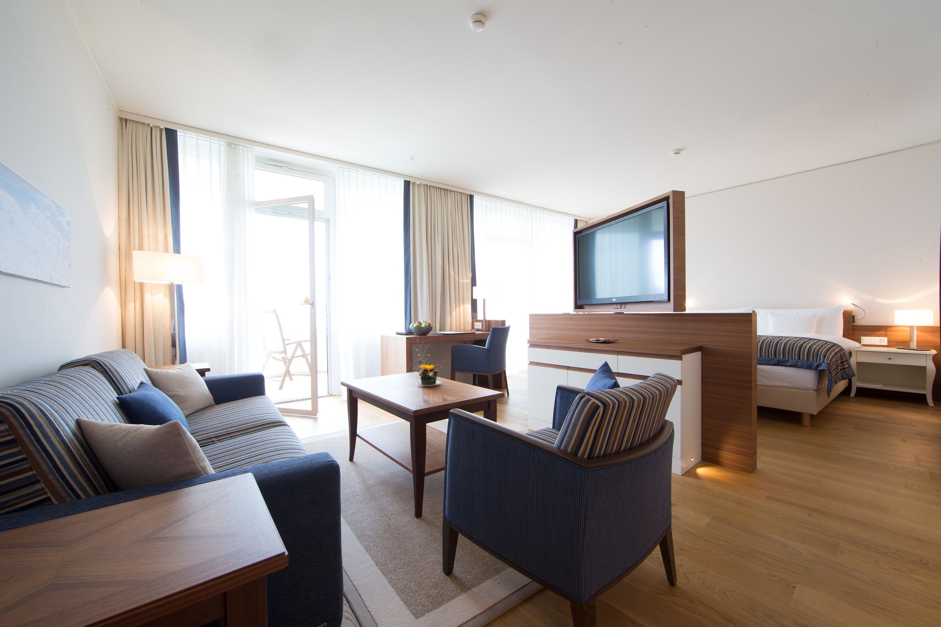 Una habitación de hotel moderna con sofá, sillón, mesa de comedor y televisor montado en la pared.