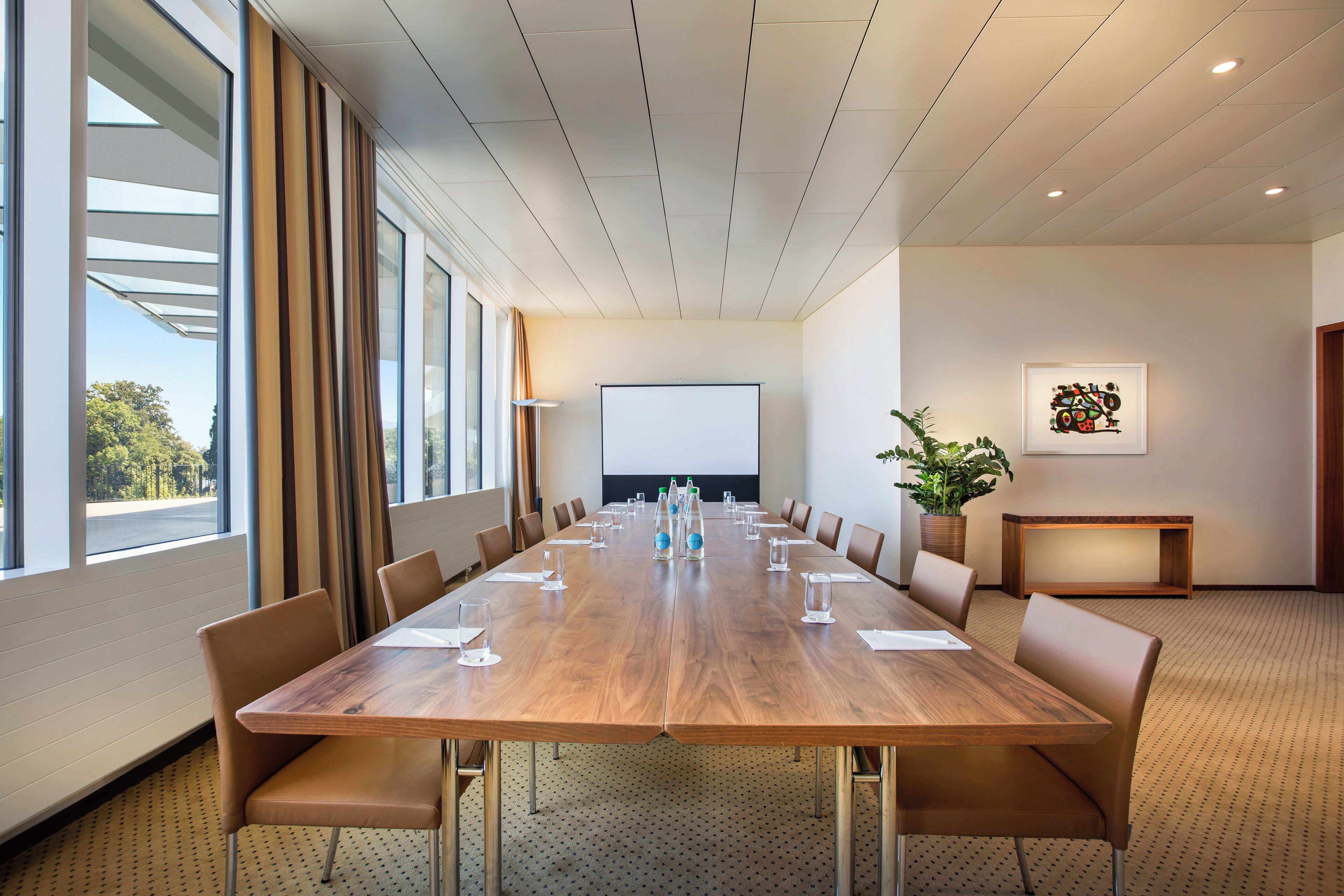 Una sala de conferencias con una larga mesa de madera, sillas de cuero marrón, una pantalla de proyector y una planta en maceta.