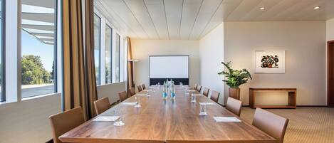 Una sala de conferencias con una larga mesa de madera, sillas de cuero marrón, una pantalla de proyector y una planta en maceta.