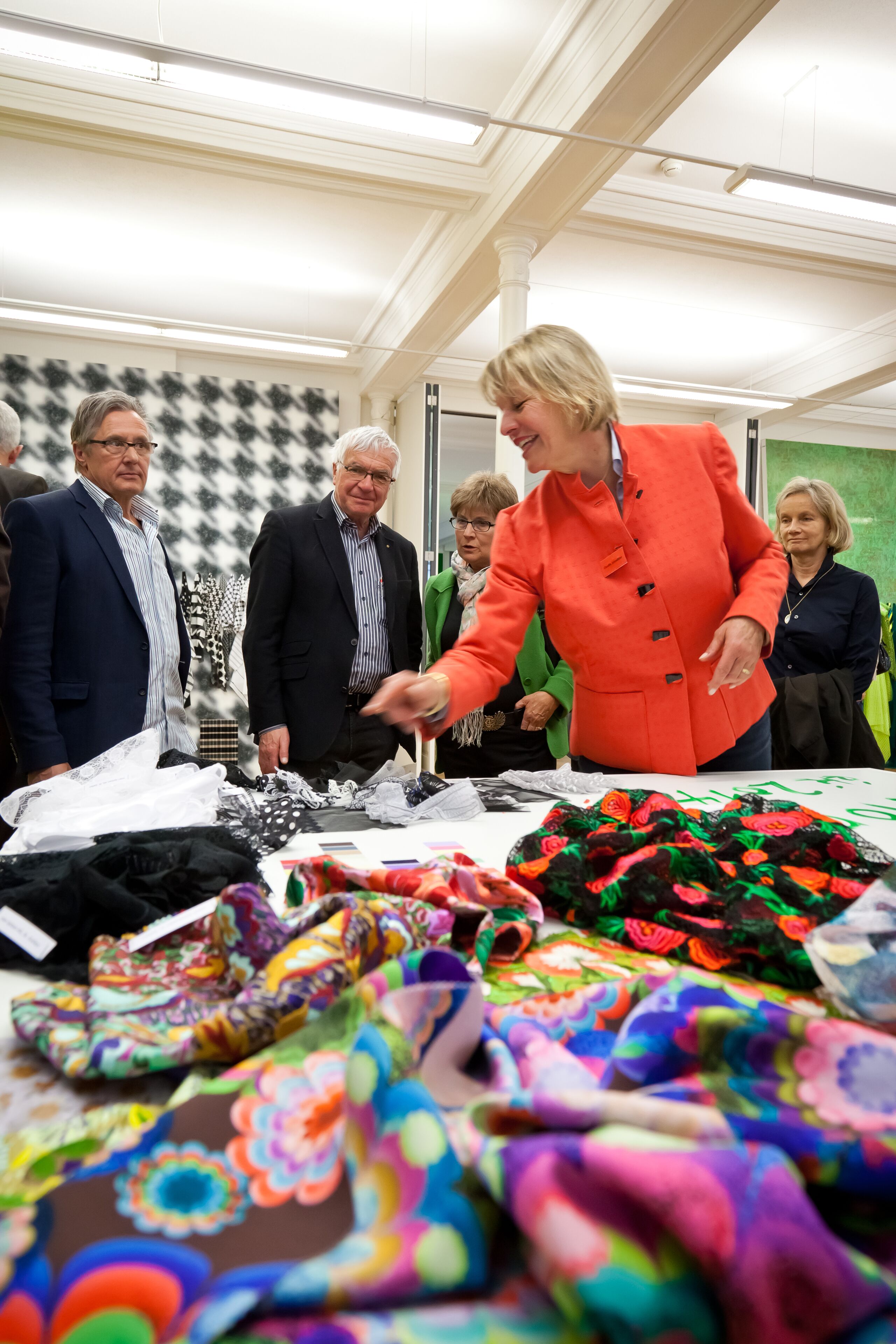 Eine Gruppe von Personen in einem Raum mit gemustertem Wandpapier und einem Tisch, auf dem farbenfrohe Stoffe ausgestellt sind.