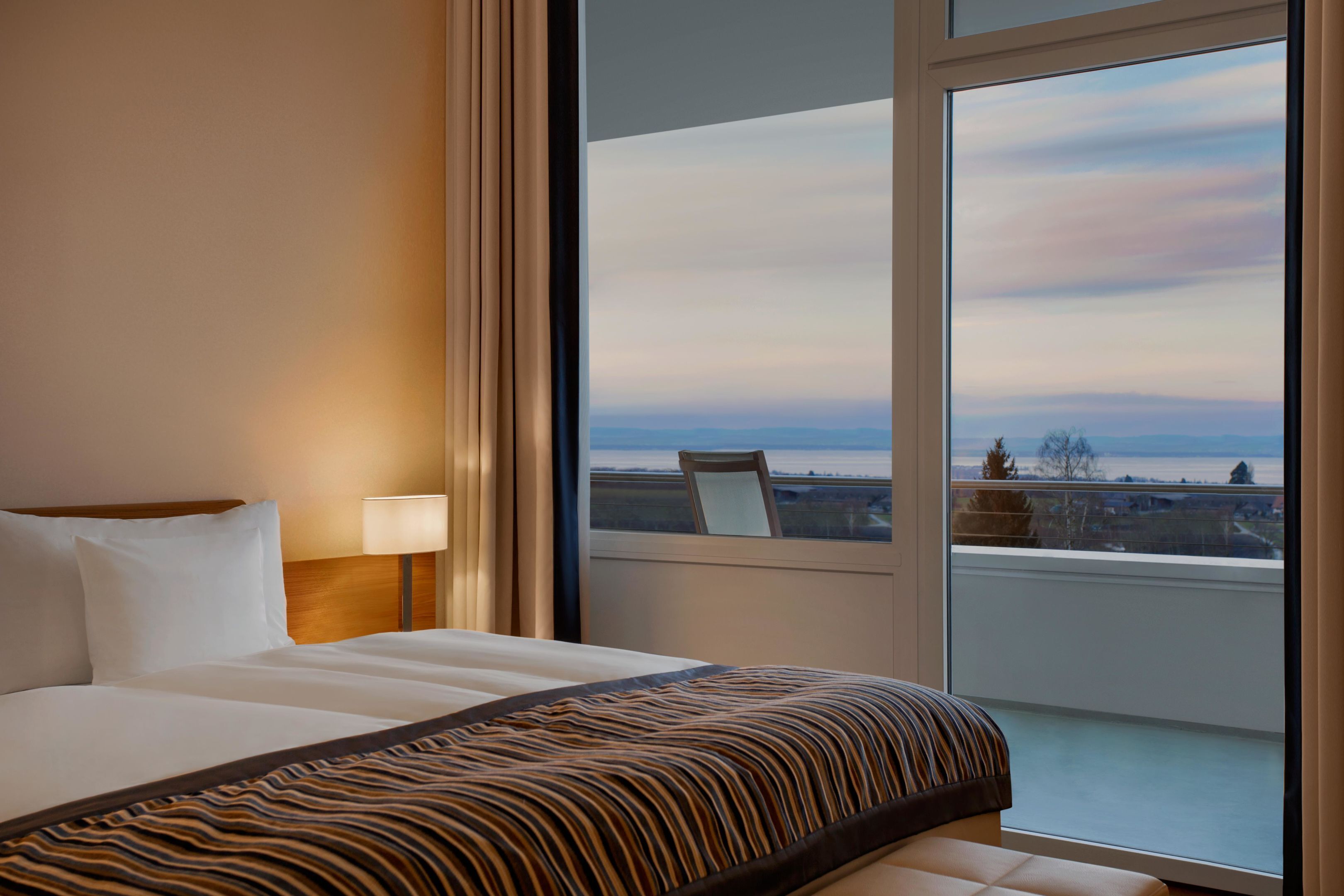Habitación de hotel con una cama grande, una mesita de noche con lámpara y vistas de la ciudad a través de un ventanal amplio.