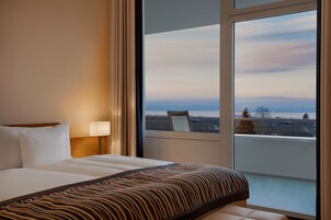 Habitación de hotel con una cama grande, una mesita de noche con lámpara y vistas de la ciudad a través de un ventanal amplio.