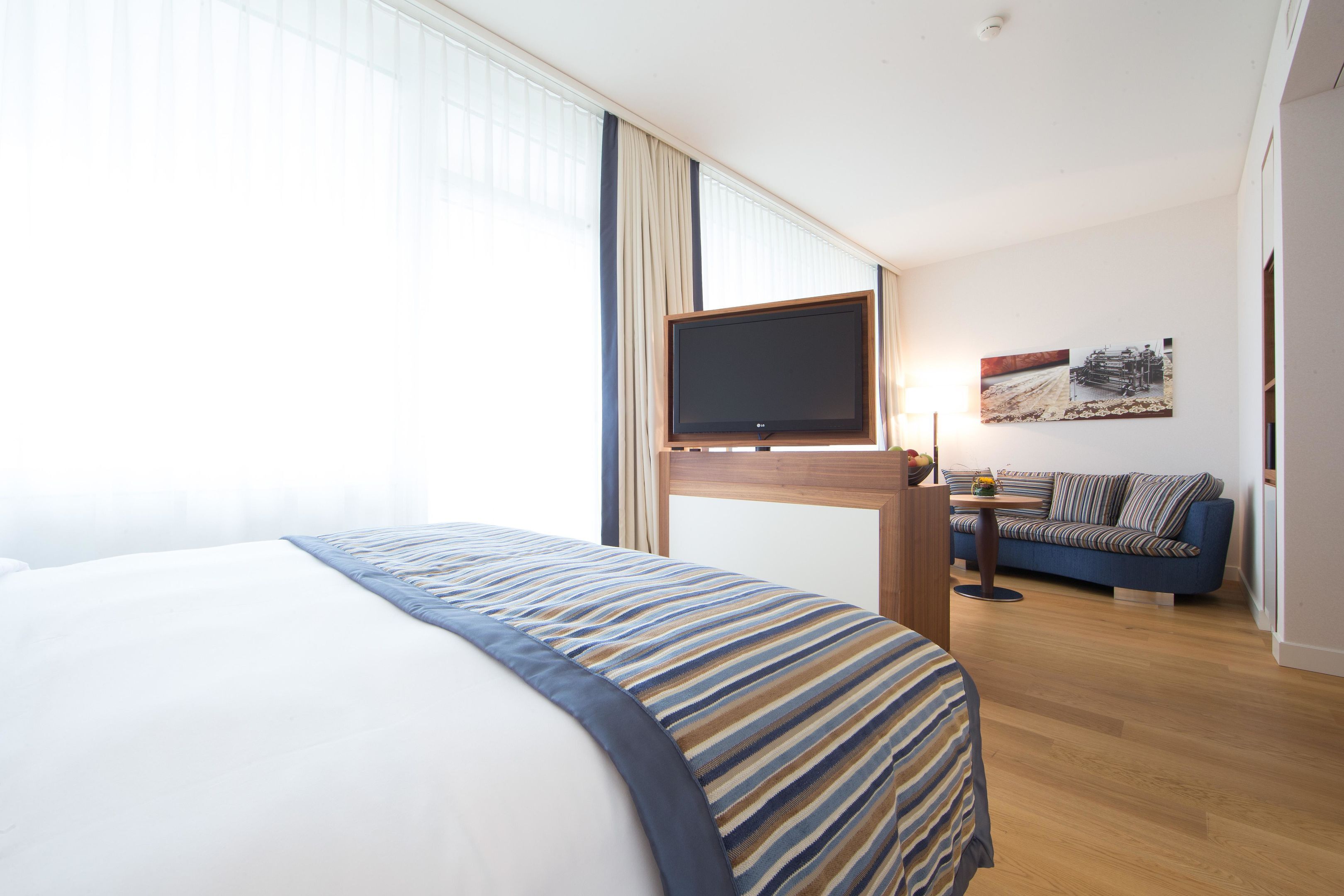A hotel room with a large bed, a TV mounted on a wooden stand, a sofa, and a painting on the wall.