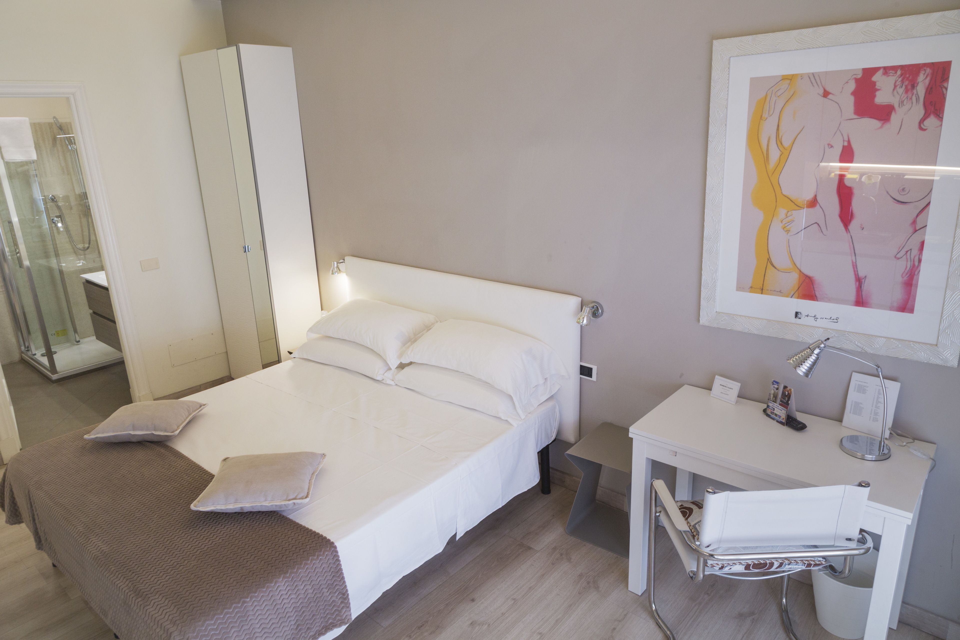 A hotel room with a bed, a desk, a chair, a wardrobe, and a bathroom visible through a glass door.