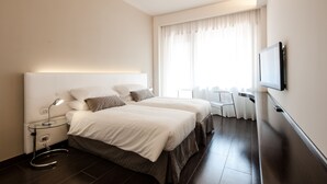 Hotelkamer met twee bedden, een aan de muur bevestigde televisie, een nachtkastje met lamp en een raam met gordijnen.