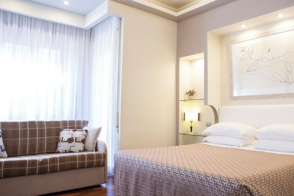 A hotel room with a bed, a sofa, a bedside table with a lamp, and a wall-mounted artwork.