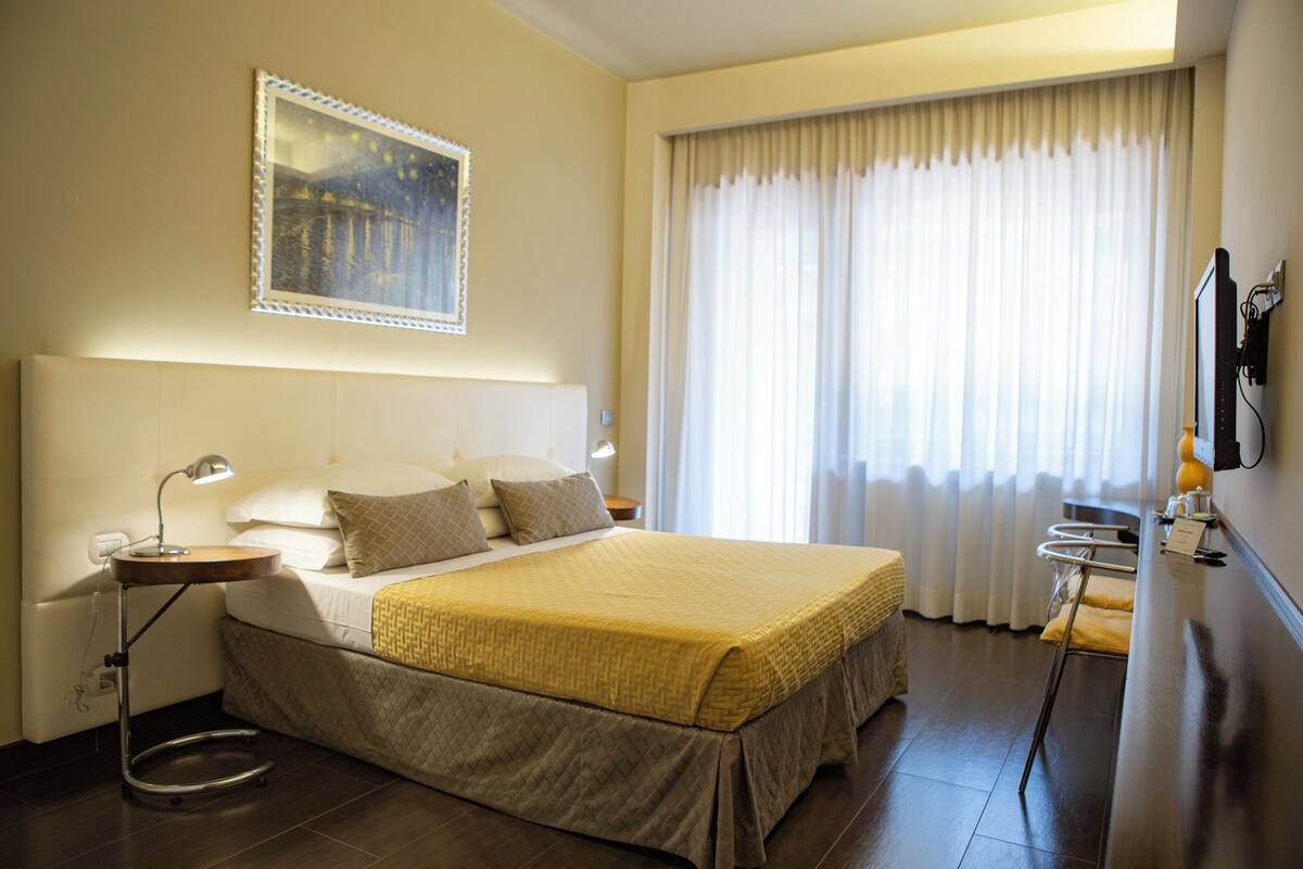 A hotel room with a large bed, bedside tables, a desk, and a window with curtains.