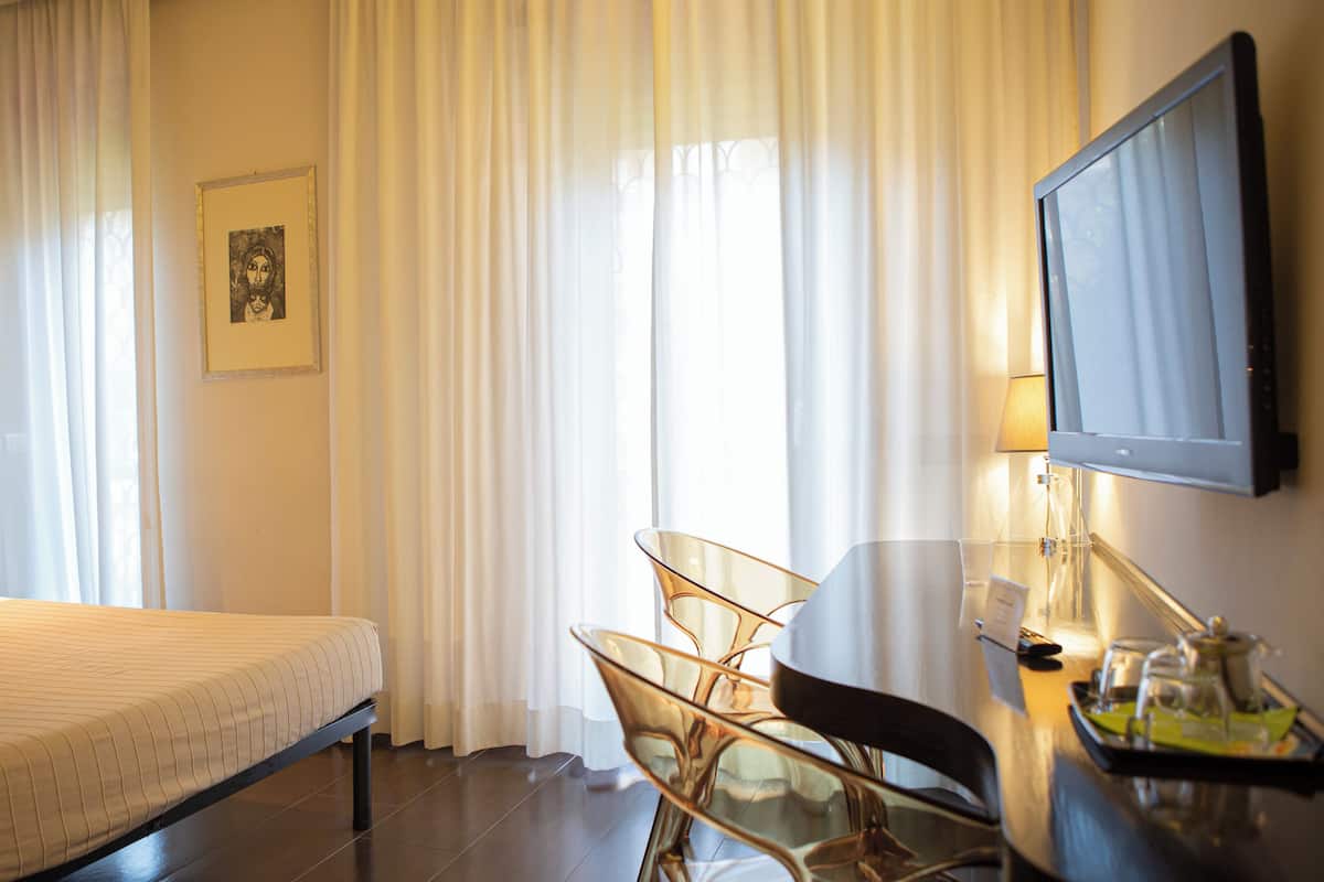 A hotel room with a bed, a desk with a chair, a TV, and a window with curtains.