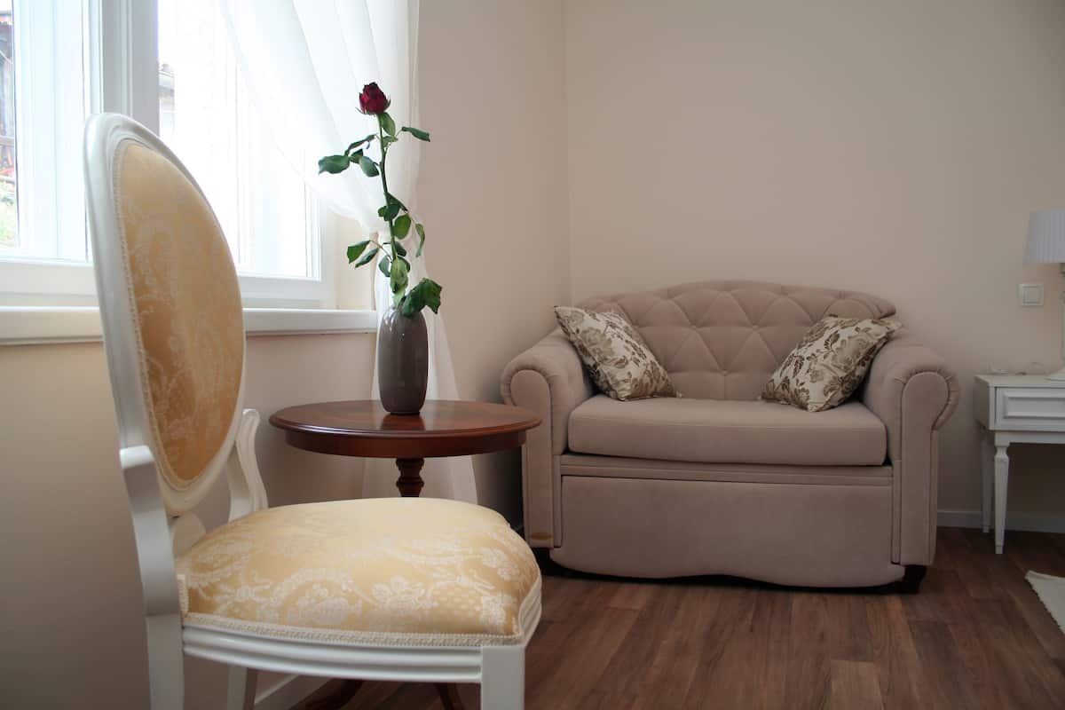 A room with a white chair, a beige sofa with floral cushions, a small round table with a vase of a single rose, and a lamp on a side table.