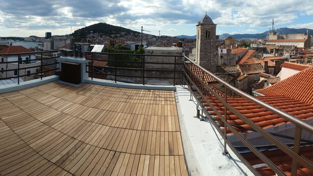 A rooftop terrace with a wooden deck, metal railings, and a view of a cityscape with buildings and a hill in the background. - Banica Apartments & Rooms (Split)