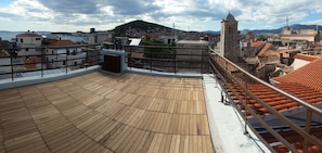 A rooftop terrace with a wooden deck, metal railings, and a view of a cityscape with buildings and a hill in the background. - Banica Apartments & Rooms (Split)