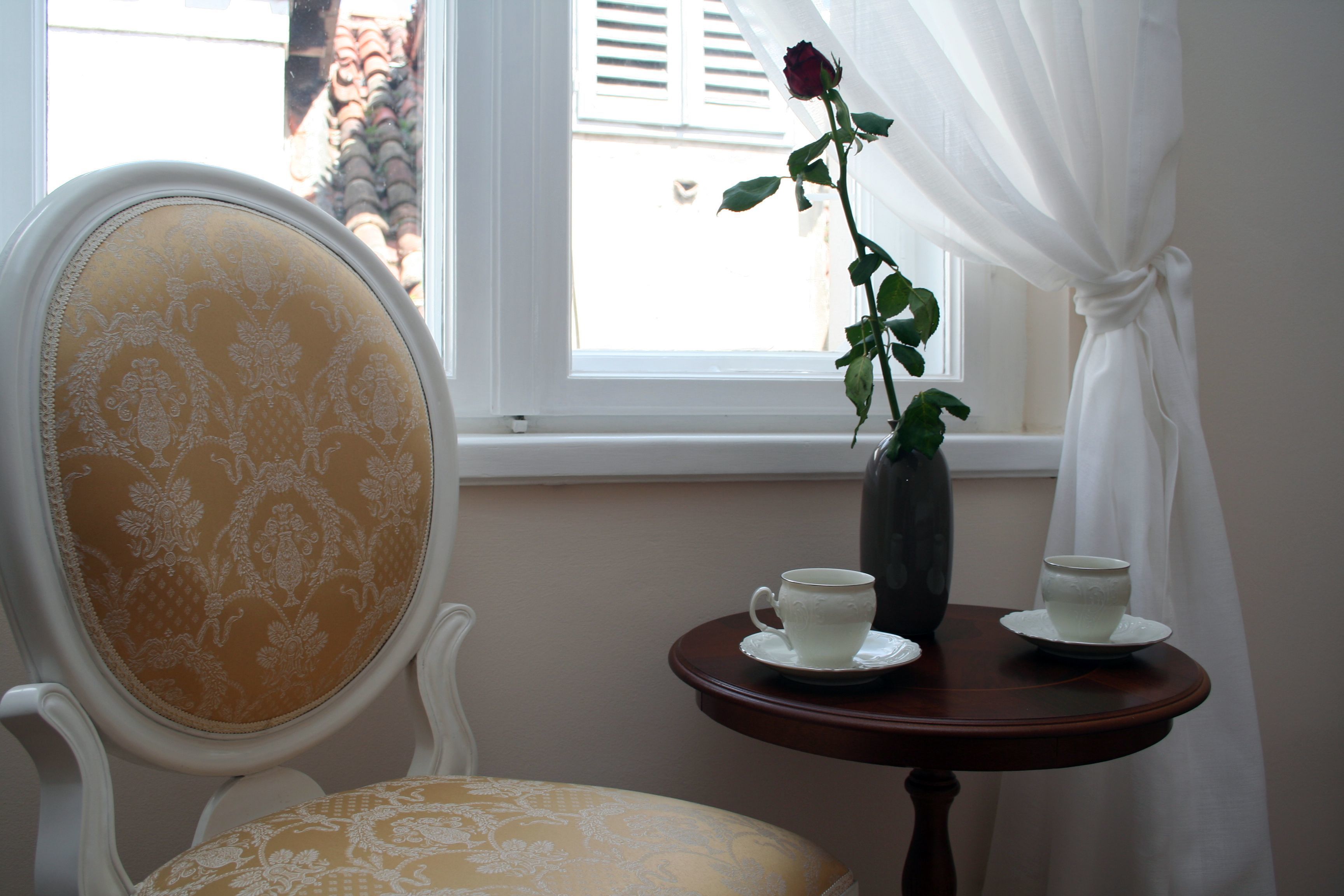 A room with a chair, a small table with a vase and two cups, and a window with curtains.
