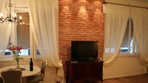 A living room with a brick wall, a television on a wooden cabinet, a dining table with chairs, and a chandelier. - Banica Apartments & Rooms (Split)