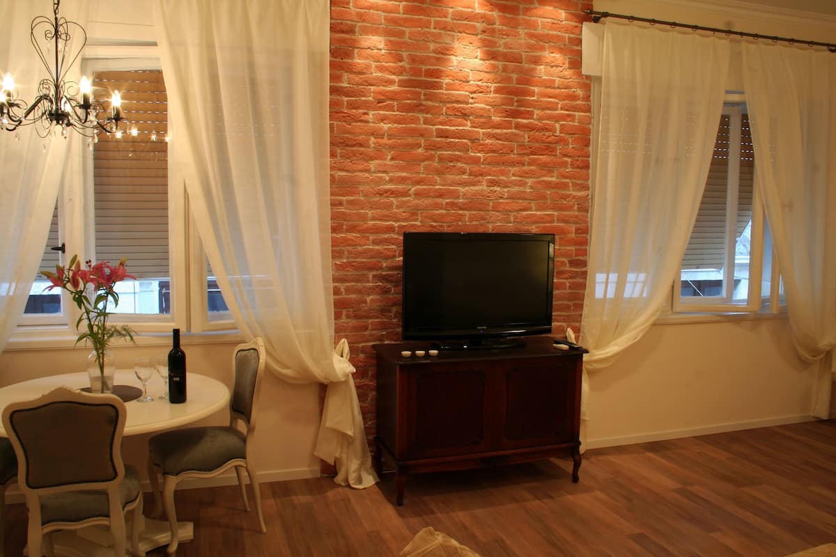 A living room with a brick wall, a television on a wooden cabinet, a dining table with chairs, and a chandelier.