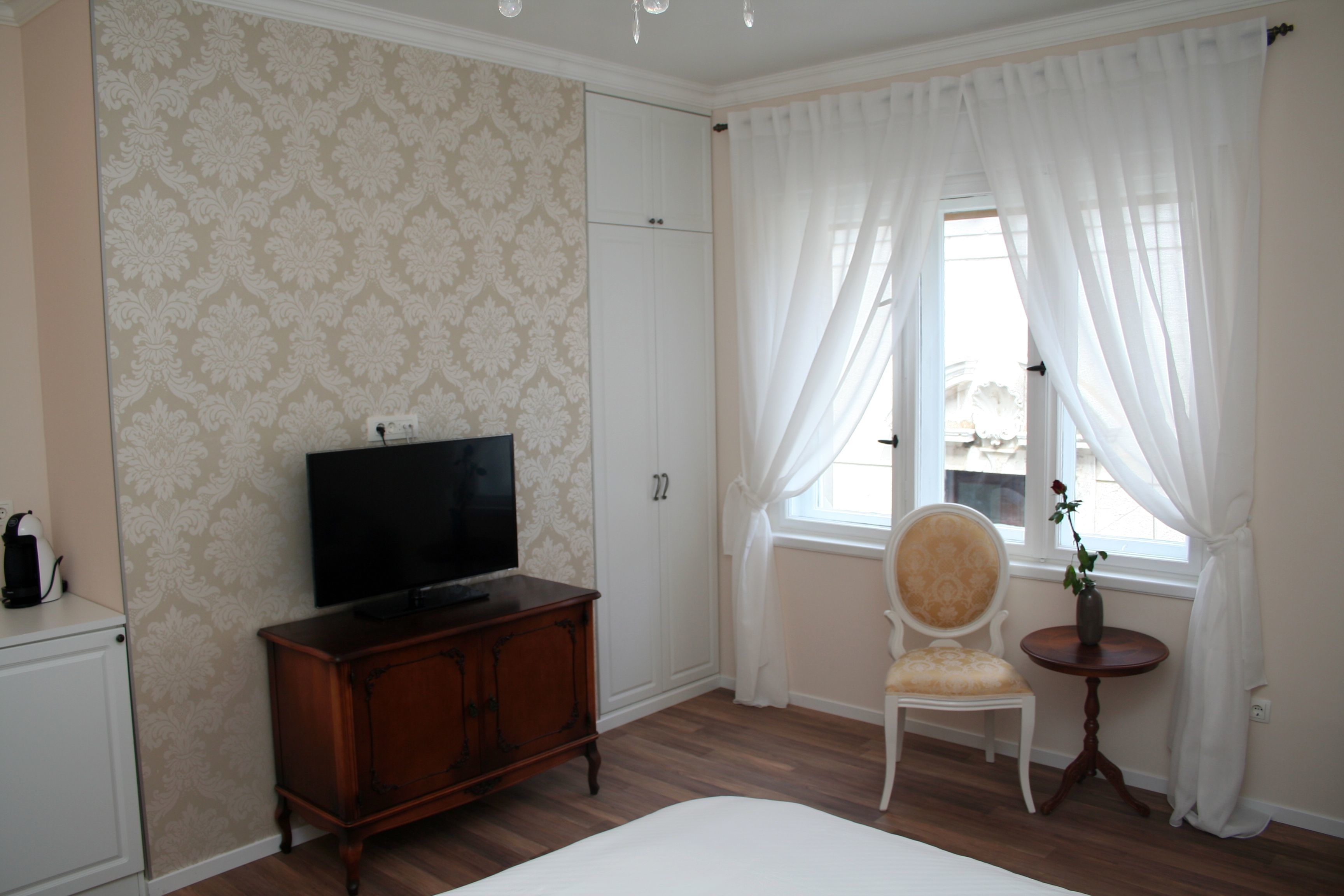 Una habitación de hotel con una cama, un televisor sobre un mueble de madera, una silla, una mesita con un jarrón y una ventana con cortinas blancas.