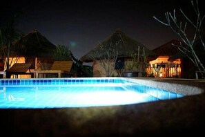Vista nocturna de una piscina con cabañas de techo de paja al fondo.