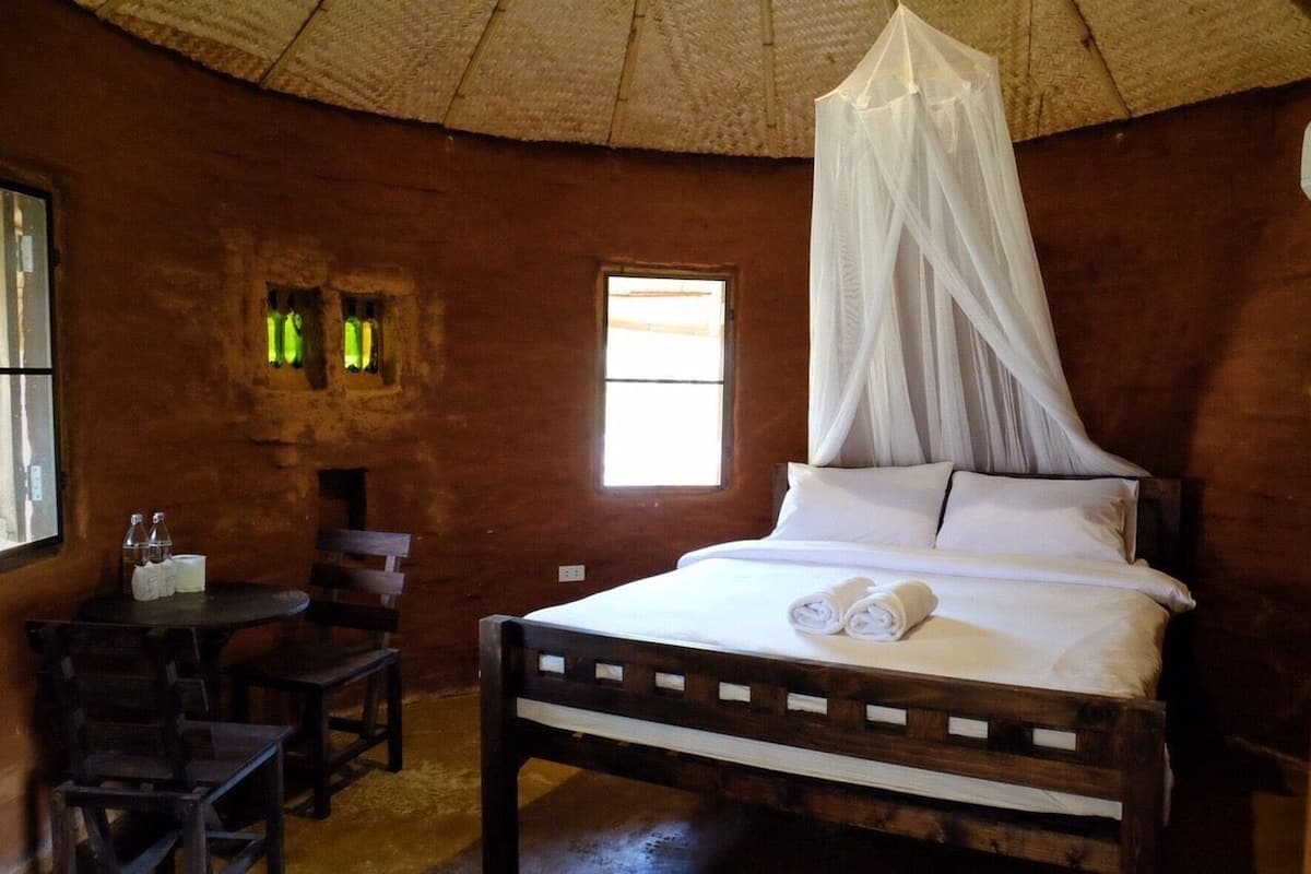 Una habitación con una cama de madera, una mesa pequeña con sillas, una ventana y un techo de paja.